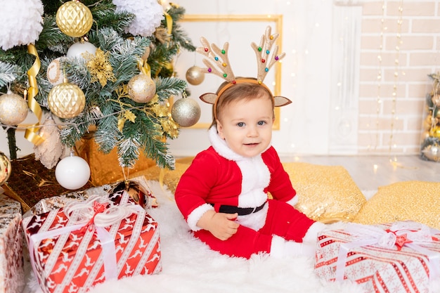 Un niño disfrazado de Santa se sienta en el árbol de Navidad con regalos, el concepto de año nuevo y Navidad