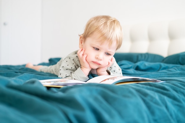 Niño divertido feliz en pijama leyendo el libro acostado en la cama de sus padres