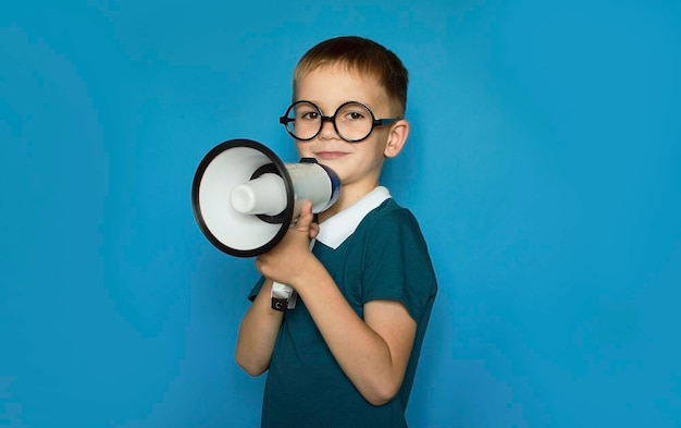 Un niño divertido con un fondo azul aislado grita por megáfono para anunciar algo que apunta hacia adelante Espacio para texto Concepto de estilo de vida infantil