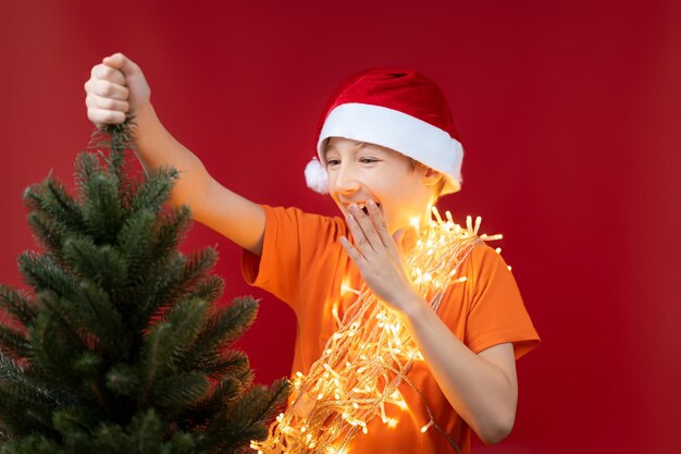 Un niño divertido con un gorro de Papá Noel sostiene la parte superior del árbol de Navidad y se ríe alegremente