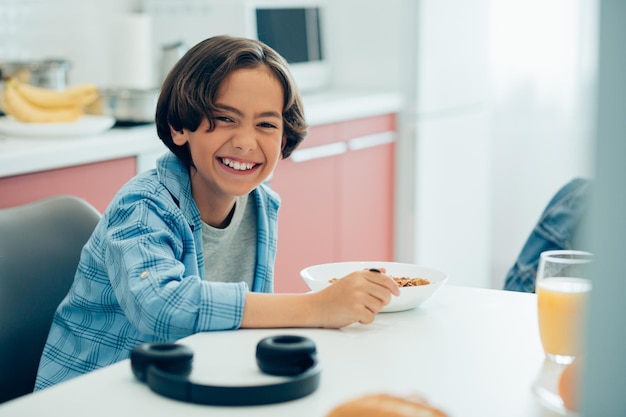 Foto niño divertido sentado en la mesa de la cocina con una cuchara y un plato de cereales niño mostrando sus dientes mientras sonríe