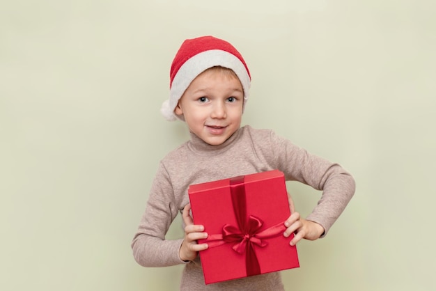 Un niño divertido con un sombrero de Santa Claus sostiene una caja de regalo roja sobre un fondo claro El concepto de regalos de Año Nuevo y Navidad Copiar espacio