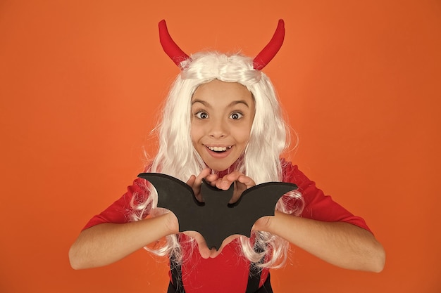 Niño divertido en sombrero de traje de bruja para Halloween con hors de diablo y peluca sosteniendo murciélago sobre fondo naranja monstruos de halloween