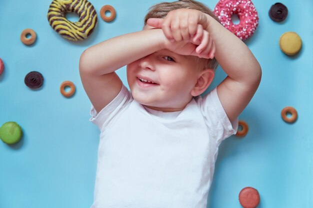 Niño divertido sonriente con macarons franceses y donas sobre fondo azul Concepto del Día Nacional de la Dona Vista superior Foto de alta calidad