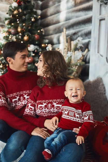 Niño divertido con un suéter feo posando con sus padres en el interior de Navidad. Los padres jóvenes y su pequeño hijo en ropa de Navidad