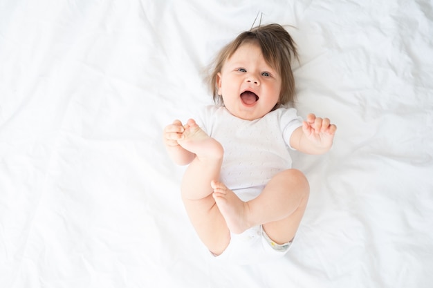 Niño divertido en traje blanco sonriendo y acostado en una ropa de cama blanca en casa. vista superior.