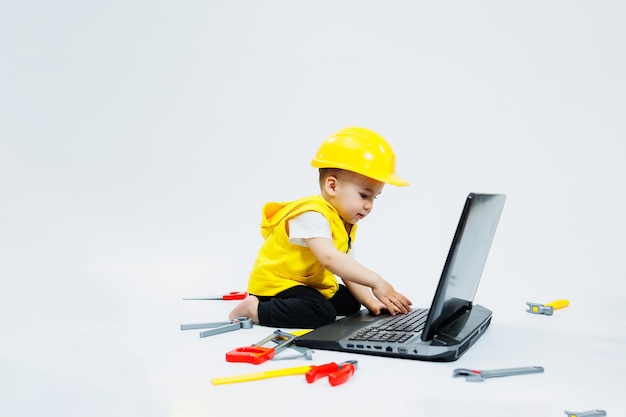 Un niño de dos años con un chaleco amarillo de fondo blanco se sienta con un portátil y juega con herramientas de construcción de plástico Juguetes para niños pequeños