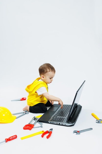 Foto un niño de dos años con un chaleco amarillo de fondo blanco se sienta con un portátil y juega con herramientas de construcción de plástico juguetes para niños pequeños