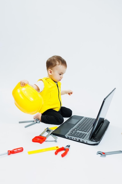 Un niño de dos años con un chaleco amarillo de fondo blanco se sienta con un portátil y juega con herramientas de construcción de plástico Juguetes para niños pequeños