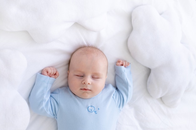 Niño duerme en la cama acostado boca arriba en pijama azul con las manos en alto entre las almohadas de las nubes, un sueño recién nacido saludable