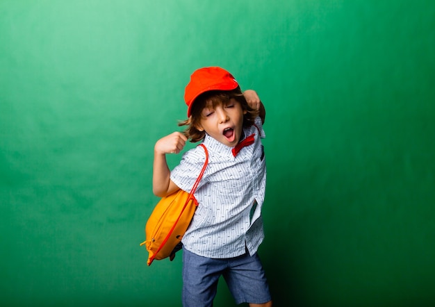 Niño emocional de 7 años con gorra roja y mochila amarilla. De pie sobre un fondo verde, divirtiéndose.