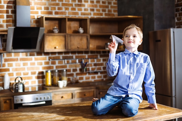 Niño encantado sentado en la cocina mientras juega con el avión de papel