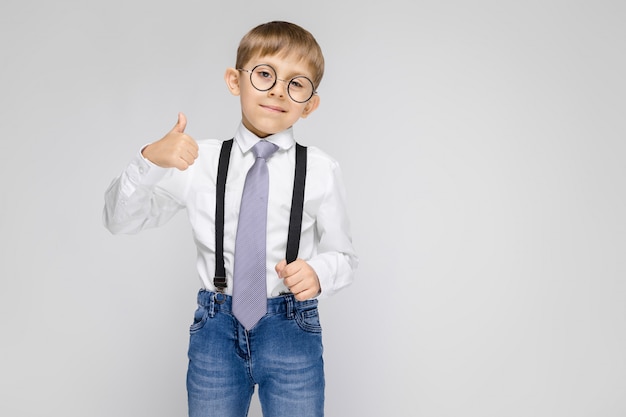 Un niño encantador con una camisa blanca, tirantes, corbata y jeans claros se encuentra en una pared gris.