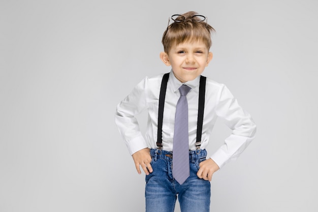 Un niño encantador con una camisa blanca, tirantes, corbata y jeans claros se encuentra en una pared gris.