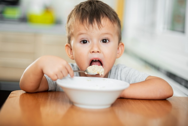 Un niño encantador con una camiseta en la cocina come avena con mucha avidez