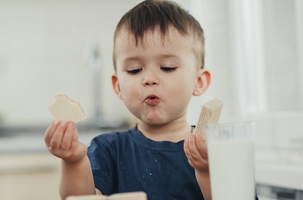 Un niño encantador come gofres en la cocina y bebe leche.