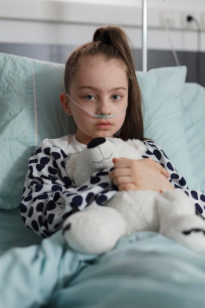 Niño enfermo que usa un tubo de oxígeno mientras está sentado en la cama del paciente con un juguete de oso de peluche. En tratamiento, una niña enferma descansa en un centro de atención médica pediátrica mientras sostiene un osito de peluche.