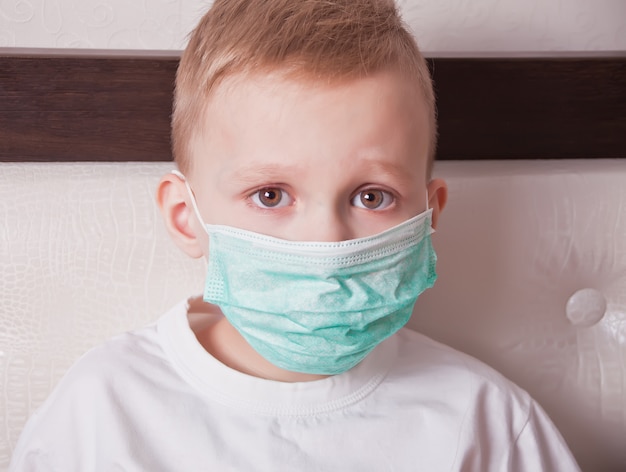 Niño enfermo sentado en la cama con máscara médica y no se siente bien
