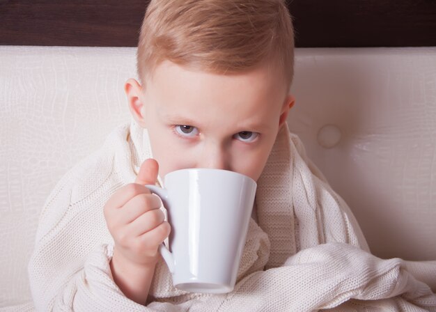 El niño enfermo sentado en una cama y sostiene una taza de té. Concepto de medicina.