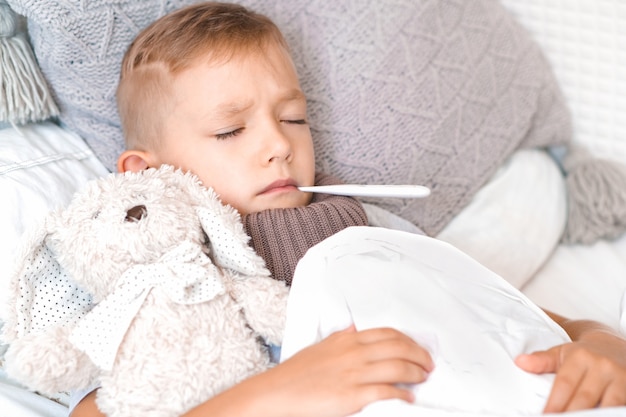 Niño enfermo y triste yace en la cama con un termómetro en la boca y un peluche en sus brazos