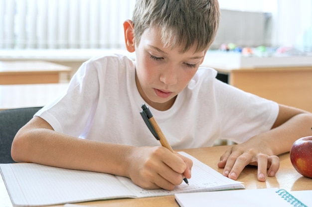 Un niño enfocado haciendo la tarea escribiendo texto en un cuaderno en la mesa Lindo niño positivo escribe y sonríe Concepto de regreso a la escuela Educación escolar y preescolar