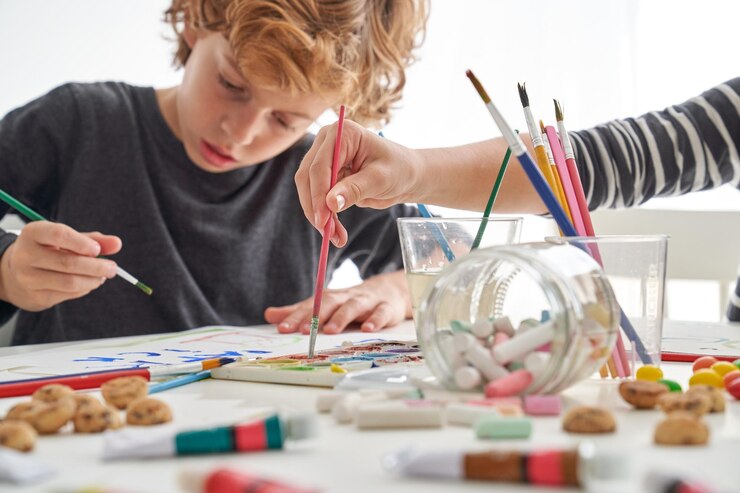 Foto niño enfocado pintando en papel con pinturas coloridas mientras está sentado en una mesa desordenada con un amigo anónimo en la sala de luz