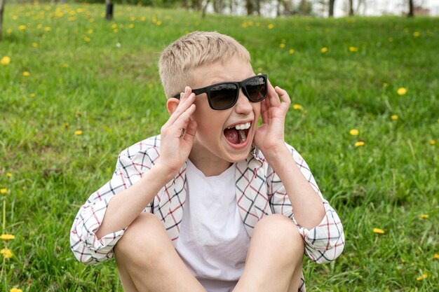 Niño enojado con gafas de sol se sienta en la hierba y grita