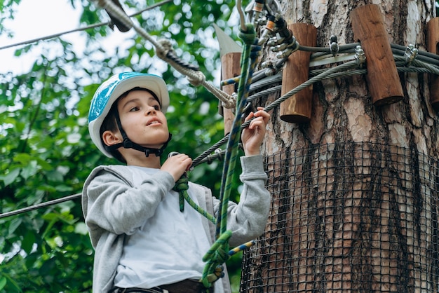 Niño en equipo deportivo, un casco protector se detuvo cerca de un árbol, sosteniendo una cuerda con las manos. Chico atractivo en un teleférico