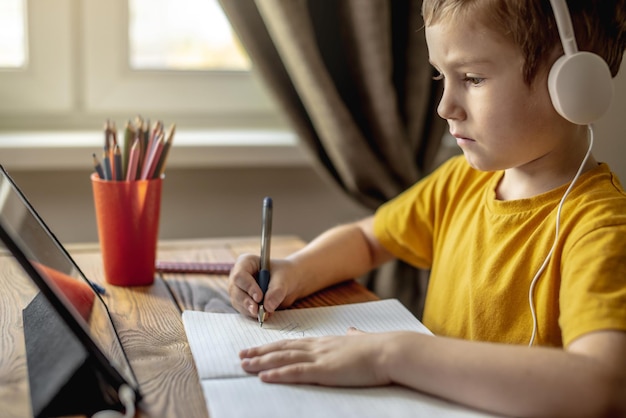 El niño es un niño con una camiseta amarilla con auriculares haciendo la tarea en una tableta Concepto de educación escolar remota