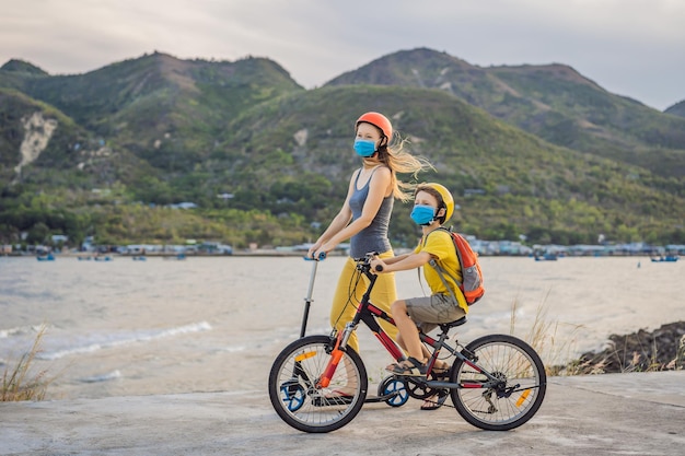 Niño escolar activo y su madre con máscara médica y casco de seguridad montando una bicicleta con mochila