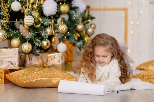 Un niño escribe una carta para Santa Claus debajo de un árbol de Navidad decorado en oro