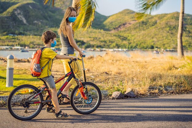 Niño de escuela activo y su madre con máscara médica montando en bicicleta con mochila en un día soleado Niño feliz en bicicleta de camino a la escuela Tienes que ir a la escuela con una máscara debido a la epidemia de coronavirus