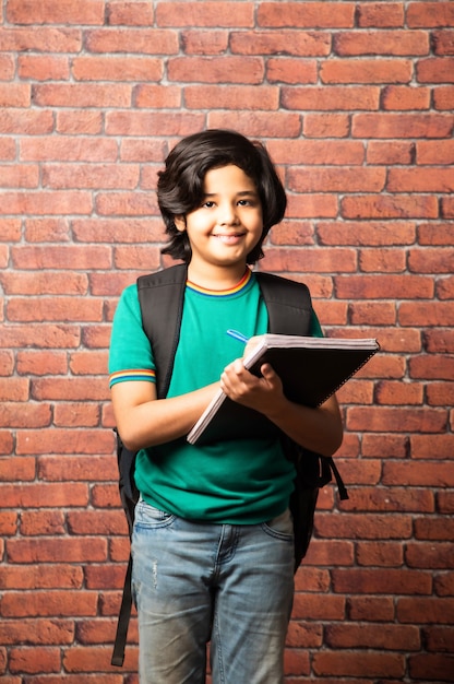 Niño de la escuela asiática india escribiendo en un cuaderno o copia con bolsa saco contra el fondo de ladrillo rojo