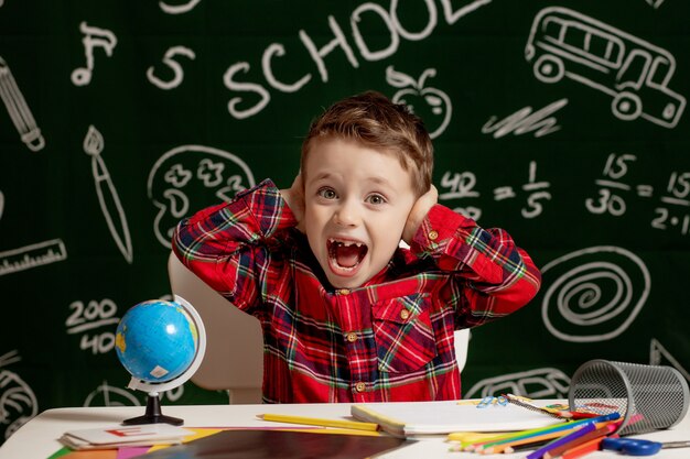 Niño de la escuela emocional sentado en el escritorio con muchos útiles escolares. Primer dia de escuela. Kid boy de la escuela primaria. De vuelta a la escuela. Niño de primaria.