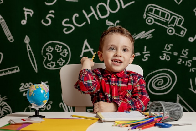 Niño de la escuela emocional sentado en el escritorio con muchos útiles escolares. Primer dia de escuela. Kid boy de la escuela primaria. De vuelta a la escuela. Niño de primaria.