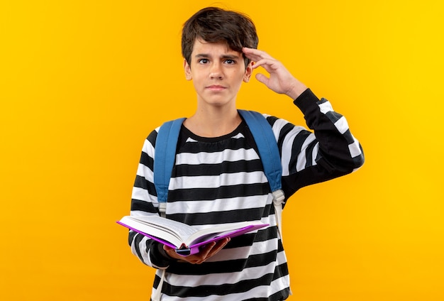 Niño de la escuela joven confidente que lleva la mochila que sostiene el libro que muestra el gesto del saludo aislado en la pared naranja