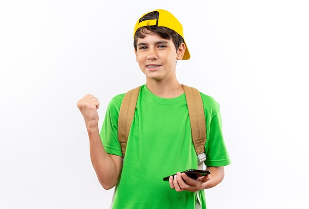 Niño de escuela joven satisfecho con mochila con gorra sosteniendo el teléfono mostrando sí gesto aislado en la pared blanca