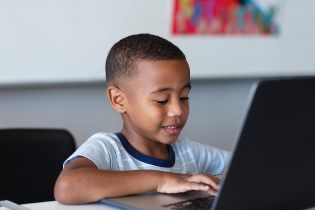 Niño de escuela primaria afroamericano sonriente usando una computadora portátil mientras está sentado en el escritorio en la escuela