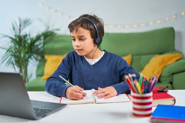 Niño de escuela que tiene clases en línea mientras está sentado en casa en cuarentena, usando la computadora portátil y tomando notas