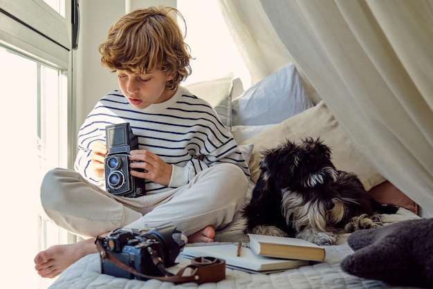 Niño de escuela sentado con las piernas cruzadas en la cama con una cámara fotográfica retro y un Schnauzer en miniatura cerca del libro en casa