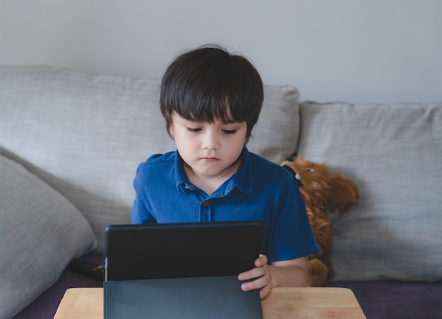 Niño de la escuela usando tableta para su tarea, Niño mirando tableta digital con cara de pensamiento, Niño viendo dibujos animados en el panel táctil, Orzuelo de nueva vida normal con aprendizaje en línea, Educación a distancia