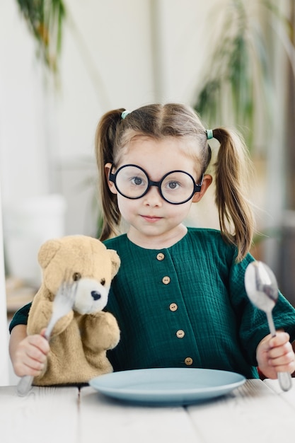 Niño esperando el desayuno con oso de juguete Niña linda en la mesa de comedor blanca en la cocina