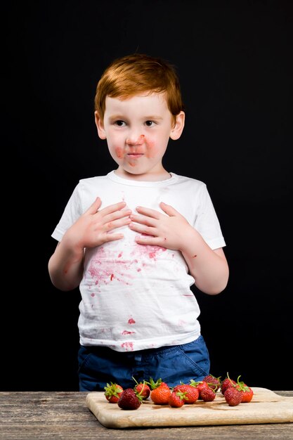 Un niño está comiendo fresas maduras y untado con bayas rojas un retrato de primer plano de un niño mientras come un postre de bayas