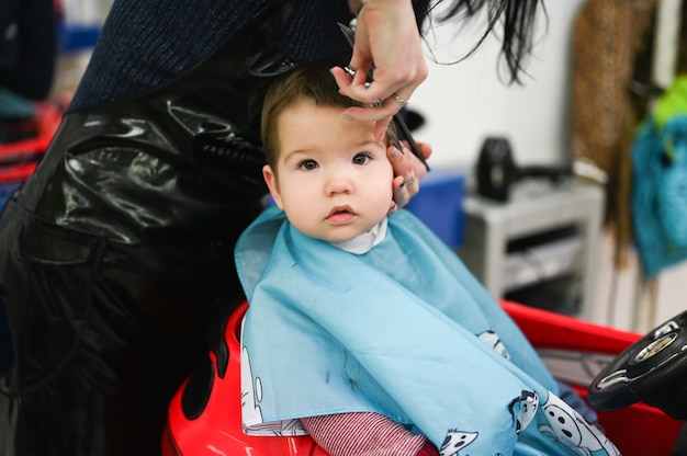 El niño se está cortando el pelo en la peluquería en un auto rojo. El primer corte de pelo del niño en la peluquería. Niño pequeño cortarse el pelo.