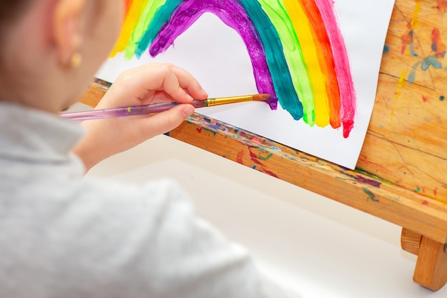 El niño está dibujando un arco iris con acuarelas en la hoja de papel del caballete. Concepto de creatividad.