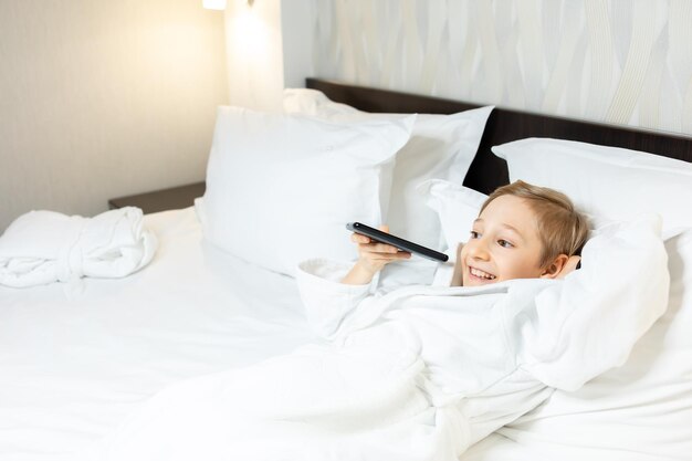 El niño está hablando por teléfono mientras está en una habitación de hotel acostado en la cama con una bata blanca.