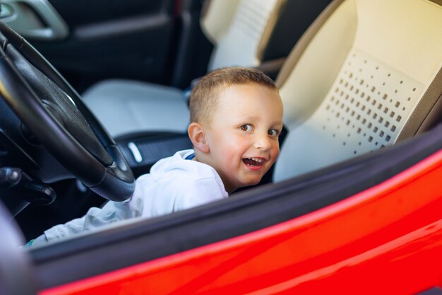 El niño está jugando en el asiento delantero del auto rojo y sonríe sinceramente