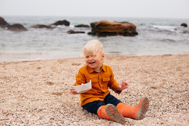Niño está jugando con un bote de papel en la playa cerca del mar Niño de vacaciones en verano u otoño en el mar con mal tiempo en otoño o verano