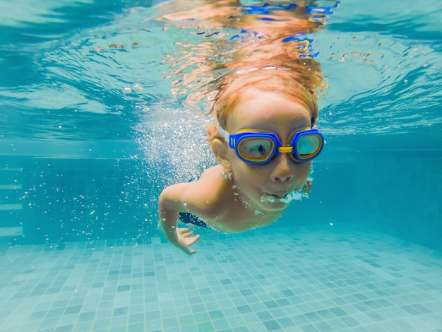 Un niño está nadando bajo el agua en una piscina sonriendo y conteniendo la respiración con gafas de natación