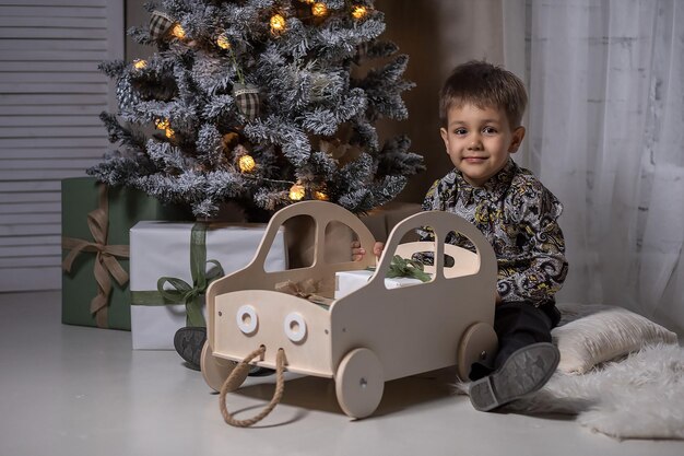 El niño está sentado en la alfombra cerca del árbol de Navidad sosteniendo un regalo y jugando con una máquina de escribir de madera